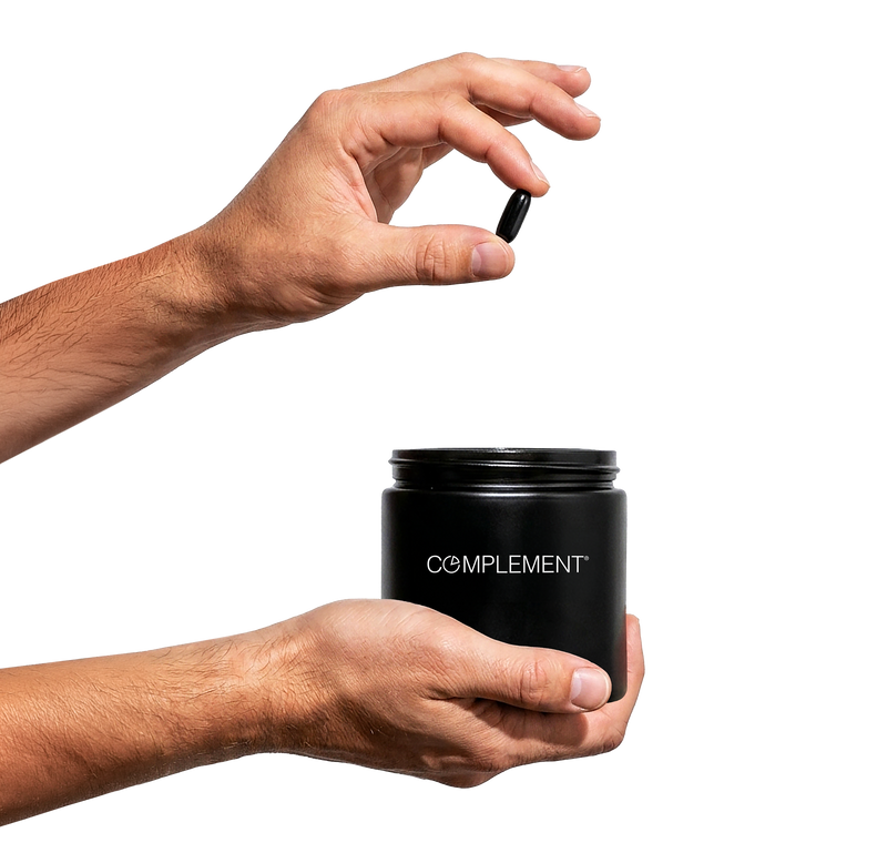 A person holds a black jar labeled COMPLEMENT in one hand and is about to drop a black capsule into the jar with the other hand, against a plain white background.