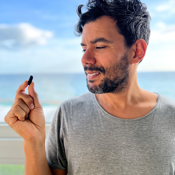 A man with dark hair and a beard, wearing a gray t-shirt, smiles while holding a small black object in his hand. He is standing outdoors with the ocean and sky in the background.