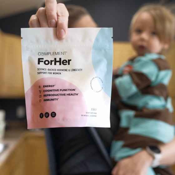 A person holds a bag of Complement ForHer supplements in focus, with a young child in their other arm and a kitchen setting in the blurred background.