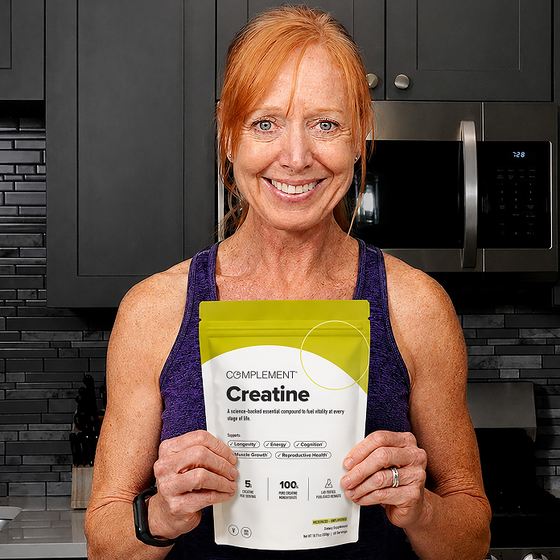 A smiling woman with red hair holds a bag of Complement Creatine supplement in a modern kitchen, wearing a purple tank top.