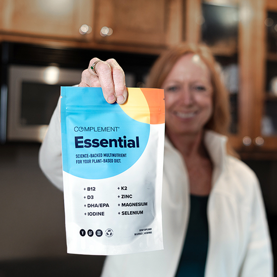 A woman stands in a kitchen smiling and holding a packet labeled Complement Essential, which lists nutrients like B12, D3, DHA/EPA, Iodine, K2, Zinc, Magnesium, and Selenium on the front.