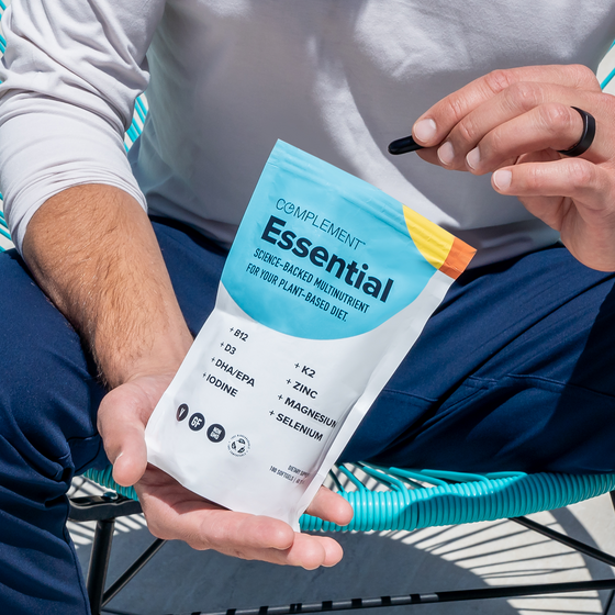 A person sits on a striped outdoor chair, holding a packet of Complement Essential multivitamins and a black capsule in their hand. The packet lists nutrients like B12, D3, K2, DHA/EPA, zinc, magnesium, iodine, and selenium.