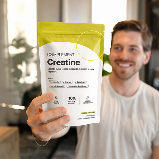A smiling person in a white shirt holds up a bag of Complement Creatine supplement, displaying the product clearly to the camera in a bright indoor setting.