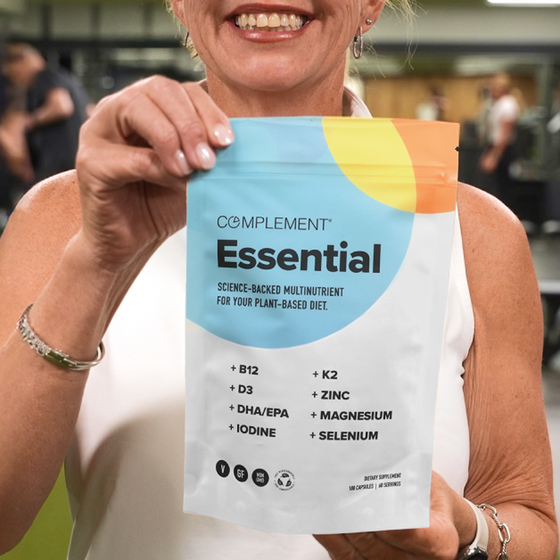 A smiling woman holds a pouch labeled Complement Essential, a science-backed multinutrient for plant-based diets, listing nutrients like B12, D3, DHA/EPA, iodine, K2, zinc, magnesium, and selenium.