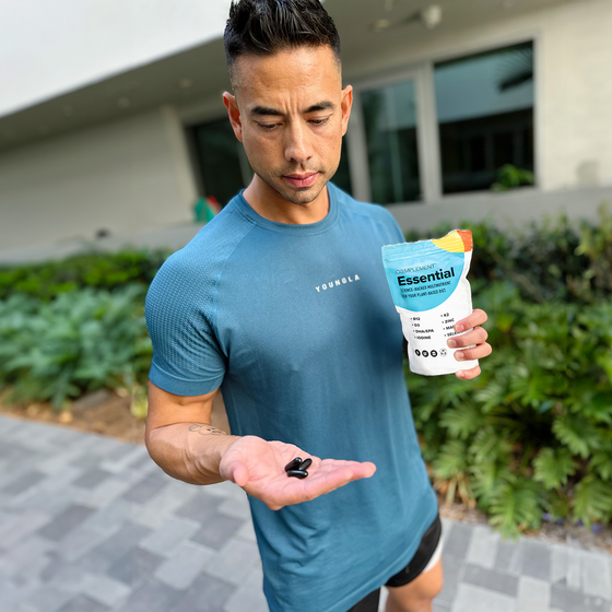 A man in a blue shirt stands outdoors holding a packet labeled Essential in one hand and shows several black capsules in his other hand, with greenery and a building in the background.