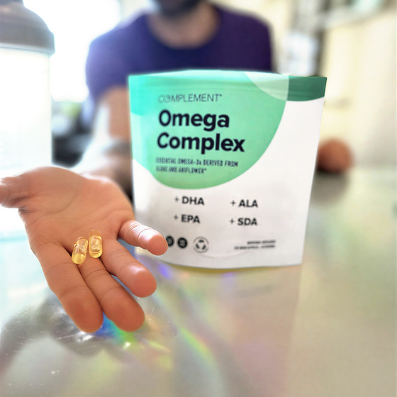 A hand holding three yellow capsules in front of a Complement Omega Complex supplement package on a table. The background is blurred, focusing on the capsules and package.