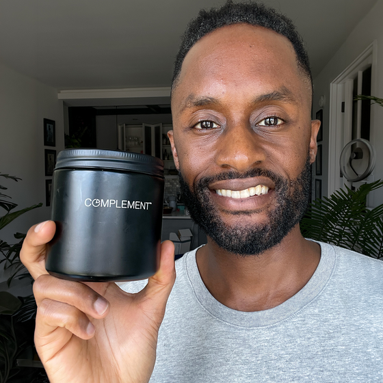 A smiling person in a gray shirt holds up a black jar labeled COMPLEMENT in a bright, modern indoor setting with plants in the background.
