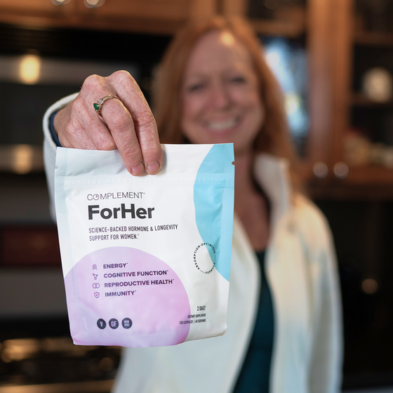 A person holds a packet labeled Complement ForHer with benefits like energy, cognitive function, reproductive health, and immunity listed. The background is softly blurred, showing another person and indoor lighting.