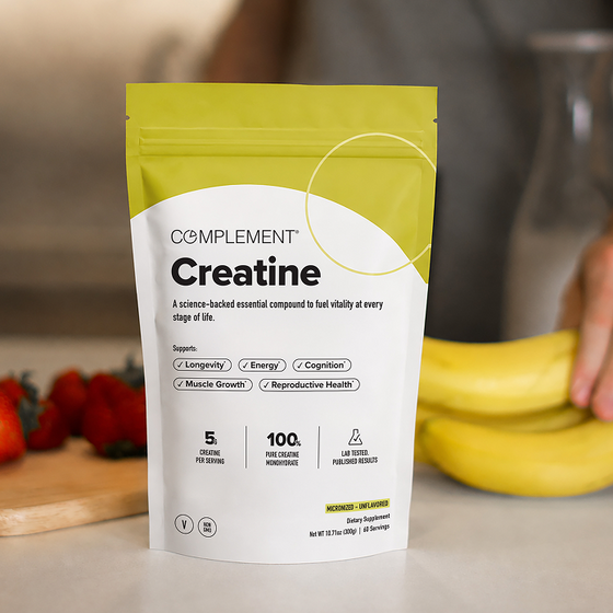 A hand holds bananas near a package of Complement Creatine powder on a kitchen counter, with strawberries and a knife in the background.