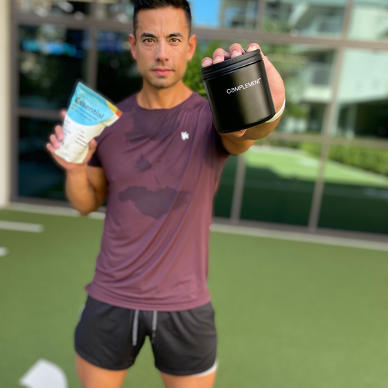 A man in athletic wear stands outdoors, holding a black container labeled COMPLEMENT in one hand and a bag of plant-based protein powder in the other. He has a sweat-stained purple shirt and is looking at the camera.