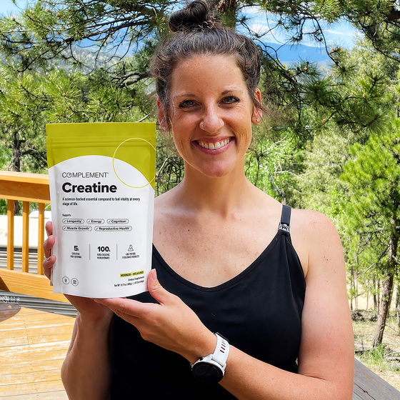 Smiling woman with brown hair in a ponytail, wearing a black tank top and white watch, holds a bag of Complement Creatine supplement outside on a wooden deck with green trees in the background.