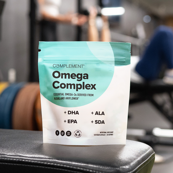 A package of Complement Omega Complex supplement stands on gym equipment, with blurred weights and a person exercising in the background. The package highlights DHA, EPA, ALA, and SDA omega-3 sources.