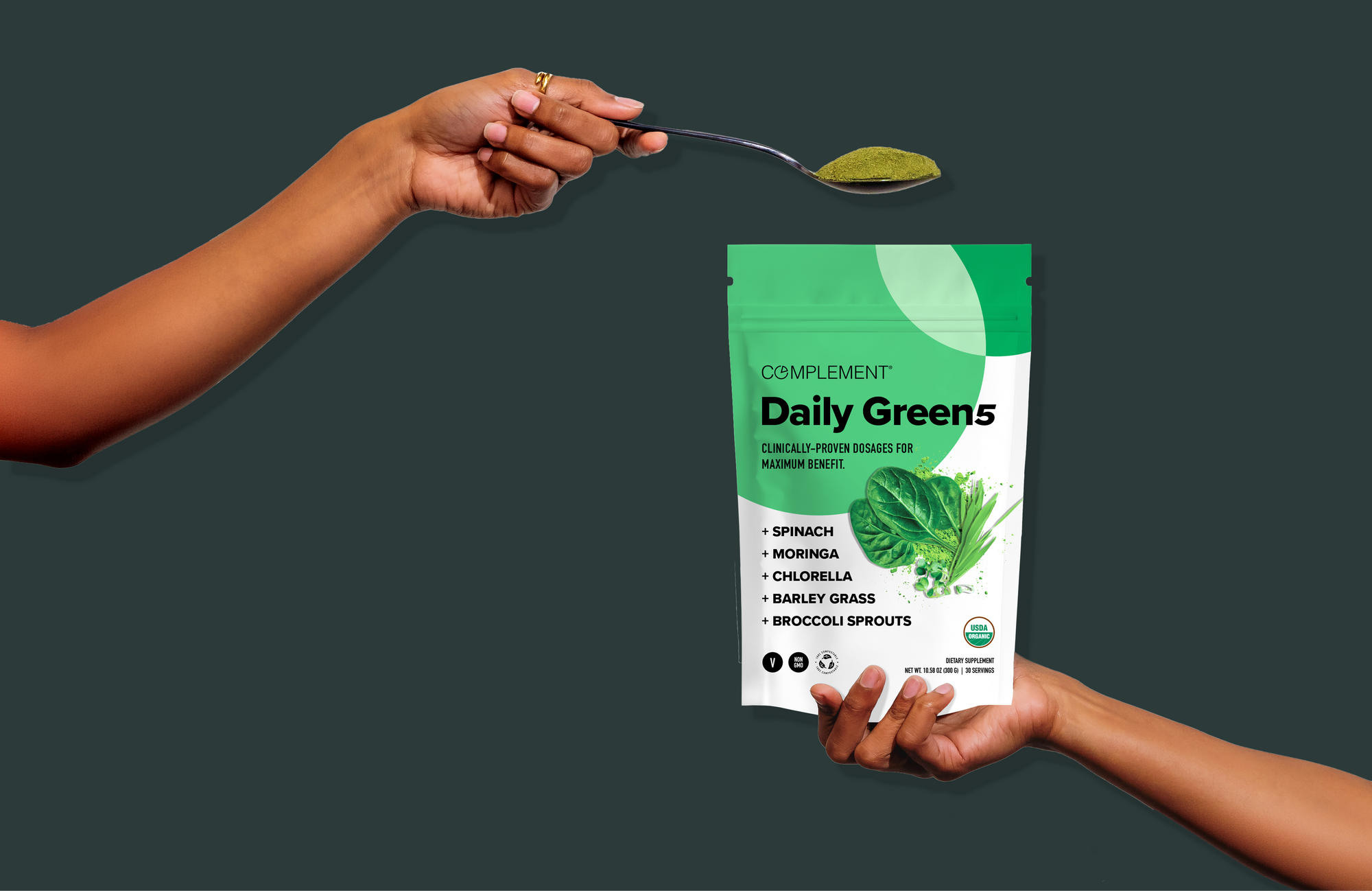 A hand holds a spoon with green powder above another hand holding a green and white bag labeled Complement Daily Greens with images of leafy vegetables and a list of ingredients.