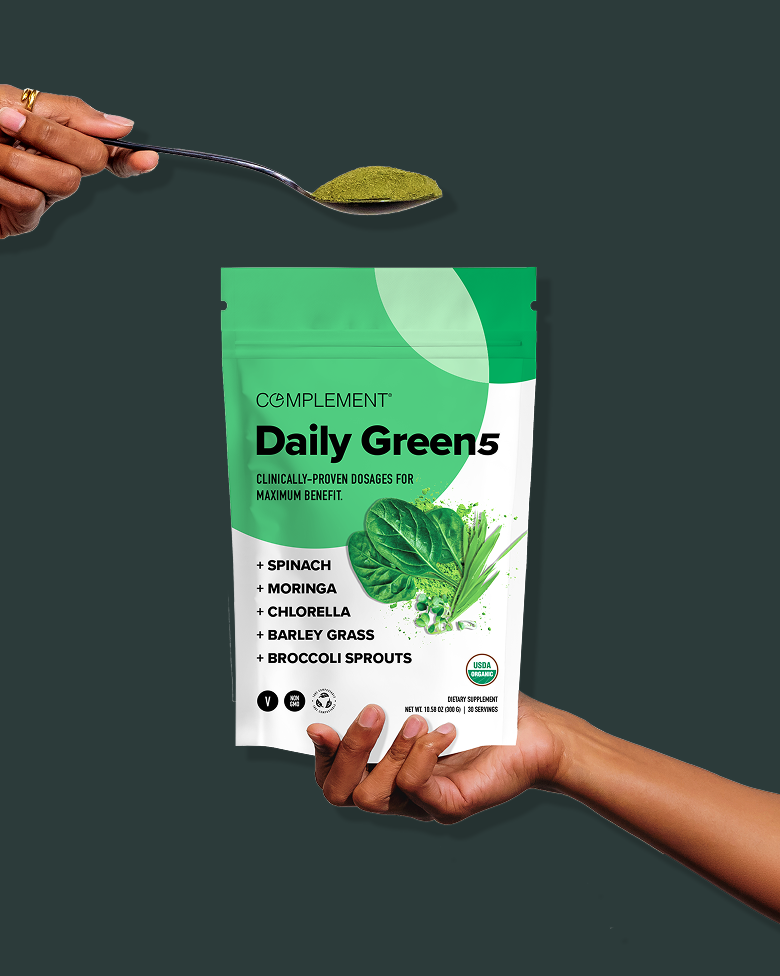 A hand holds a bag of Complement Daily Green 5 powder while another hand holds a spoonful of green powder above it. The bag lists spinach, moringa, chlorella, barley grass, and broccoli sprouts as ingredients.