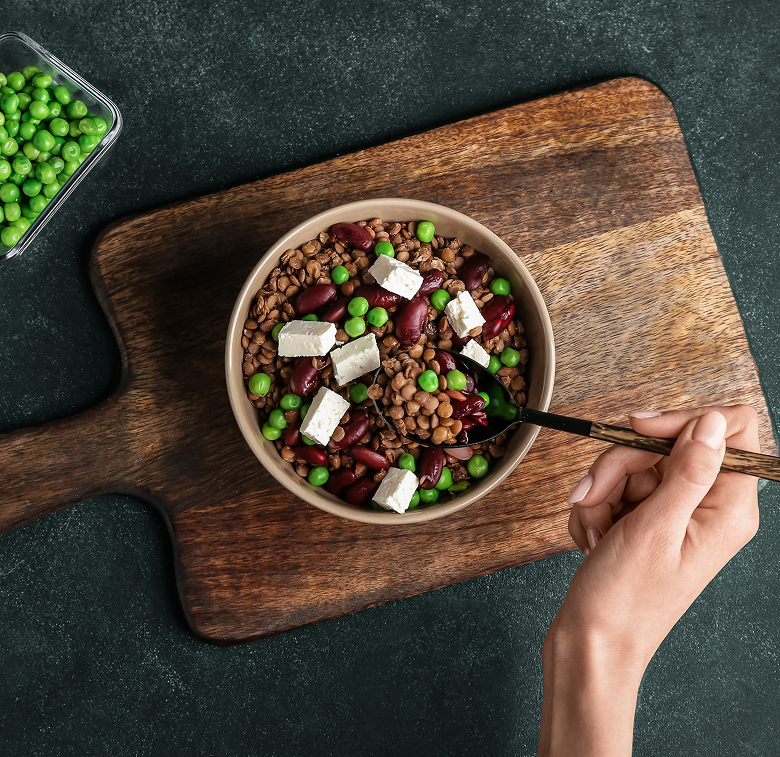A hand holding a spoon over a bowl of salad with lentils, kidney beans, green peas, and feta cheese on a wooden cutting board. A small container of green peas sits nearby.