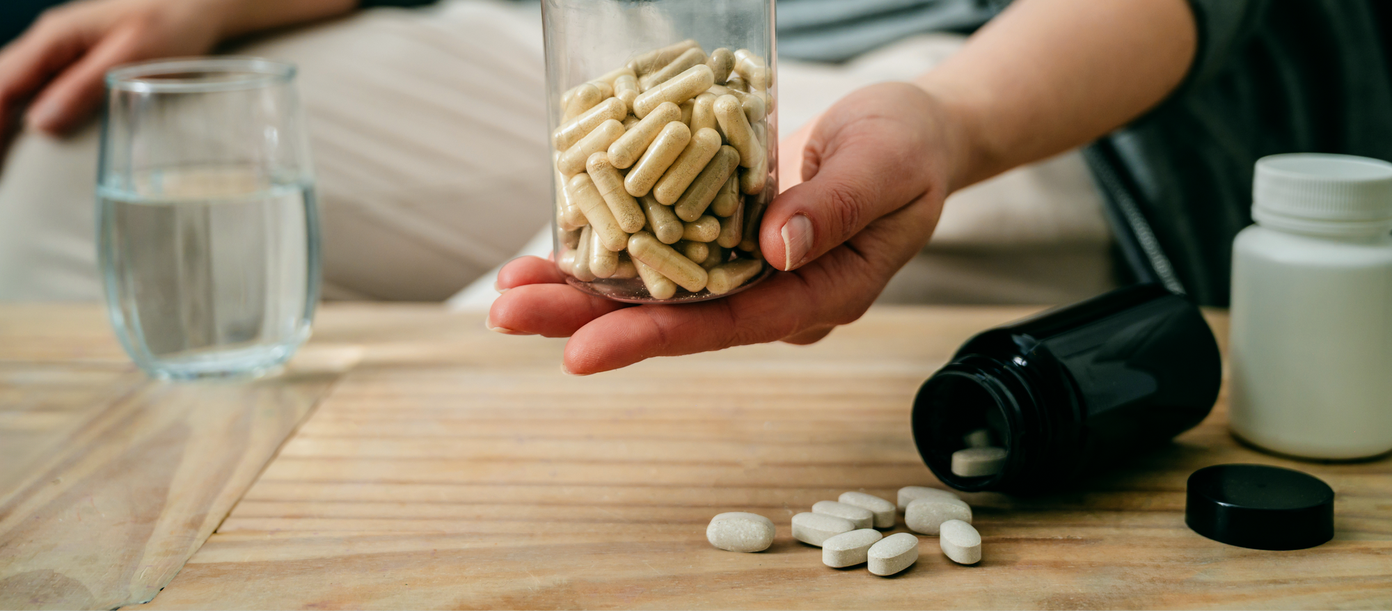 A person holds a jar filled with capsules over a wooden table with scattered tablets, a black pill bottle, a white bottle, and a glass of water in the background.