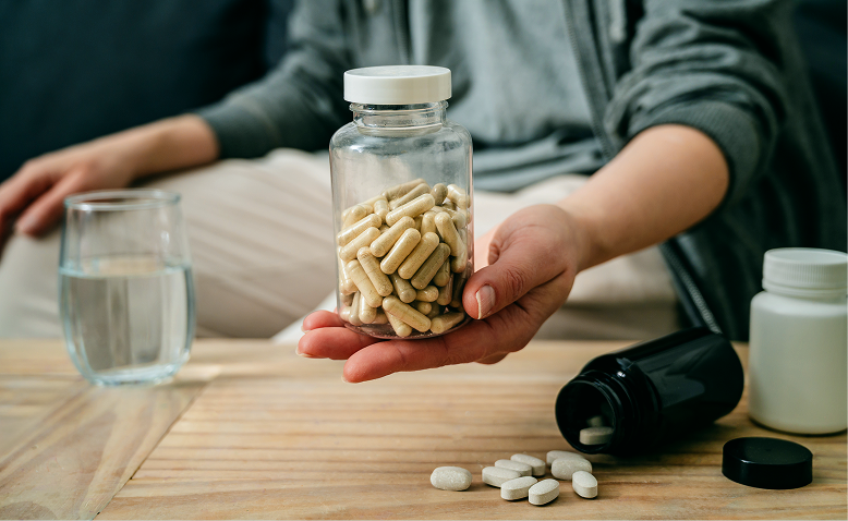 A person holding a clear bottle filled with beige capsules over a wooden table with scattered white tablets, an open black bottle, another white bottle, and a glass of water nearby.
