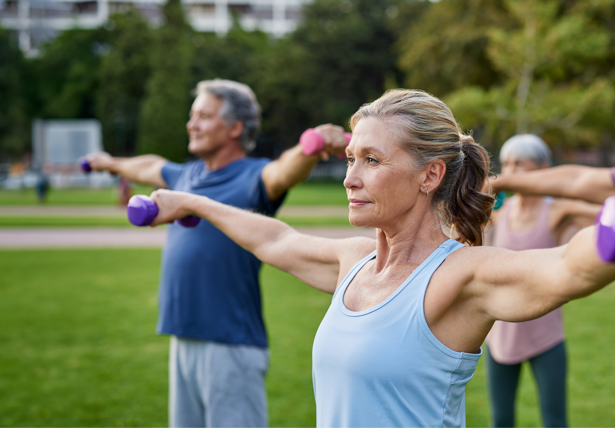 A group of older adults exercising outdoors, holding small dumbbells and raising their arms to the sides. They look focused and are standing on a grassy field with trees and buildings in the background.