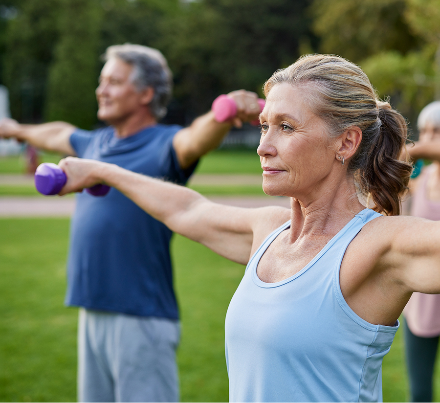 Older adults exercising outdoors, holding small dumbbells with arms outstretched to the sides. The woman in the foreground looks focused, while a man and another person are visible in the background.