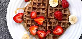 A round waffle topped with sliced strawberries and bananas, drizzled with syrup, is served on a white plate.