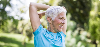 Smiling older woman with short gray hair stretches her arms behind her head outdoors, wearing a blue shirt. The background is green and blurred, suggesting a park or garden.