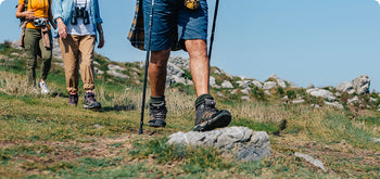 Three people hiking on a grassy, rocky trail, using walking sticks. The image focuses on their legs and feet, with hiking boots and outdoor clothing visible under a clear blue sky.