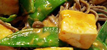 Close-up of a dish featuring snow peas, cubed tofu, and soba noodles, all coated in a glossy, orange-colored sauce.
