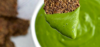 A brown, seedy cracker is being dipped into a bowl of bright green sauce or dip, with more crackers blurred in the background.