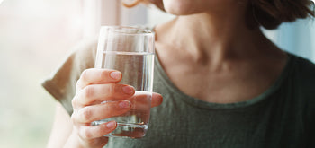 A person wearing a green shirt is holding a clear glass of water near their mouth, preparing to take a drink. The background is softly lit and out of focus.
