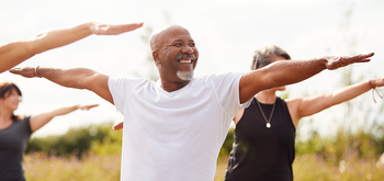 A smiling older man in a white t-shirt practices yoga outdoors with a group, all standing with arms outstretched, enjoying a sunny day in nature.