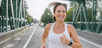 Woman wearing a white tank top and smartwatch smiles while jogging on a bridge with green railings and trees in the background.