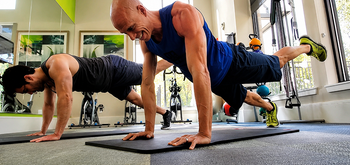 Two men in athletic wear perform plank exercises side by side on yoga mats in a bright gym, each lifting one leg off the ground. Fitness equipment and large windows are visible in the background.