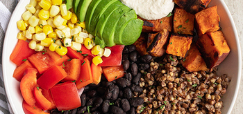 A bowl filled with sliced avocado, grilled sweet potato cubes, black beans, buckwheat, corn, chopped red bell pepper, and a dollop of creamy sauce, arranged in colorful sections.