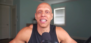 A smiling man with a bald head wearing a sleeveless black shirt speaks into a microphone in a room with light blue walls and a window with closed blinds behind him.