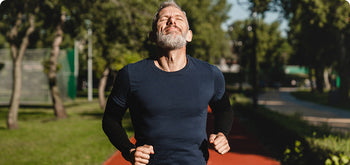 A man with gray hair and a beard runs outdoors on a red track, wearing a navy shirt and black sleeves, with trees and greenery in the background on a sunny day.
