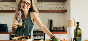 A smiling woman with long hair and glasses stands in a kitchen, holding a green smoothie. There is a salad bowl and fresh ingredients like limes on the counter in front of her.