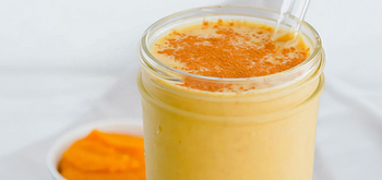 A glass jar filled with a creamy yellow smoothie, topped with a sprinkle of cinnamon, and a clear straw. In the background, there is a small bowl of orange puree.