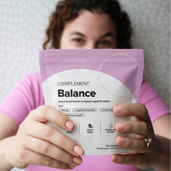 A person in a pink shirt holds up a pink and white package labeled Complement Balance, a supplement for womens health, with text highlighting energy, cognitive function, immunity, and reproductive health benefits.