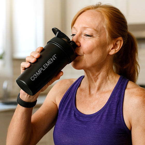 A woman in a purple tank top drinks from a black shaker bottle labeled COMPLEMENT in a bright indoor setting.