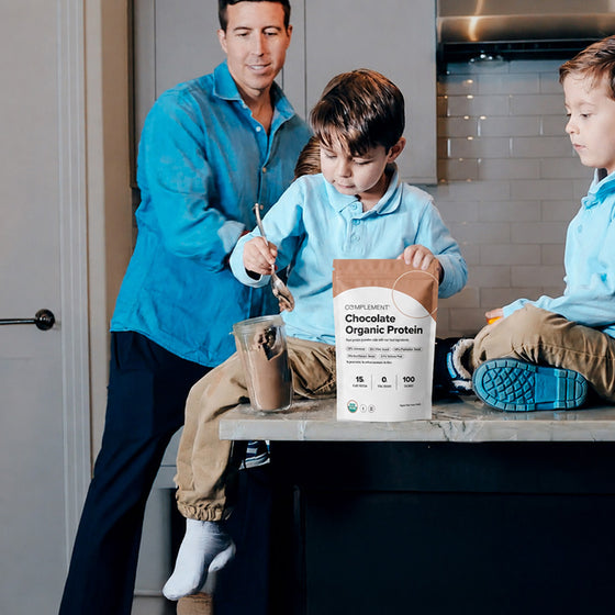 A man stands in a kitchen while two young boys in blue shirts, one sitting on a counter, make a drink with Complement Chocolate Organic Protein powder.