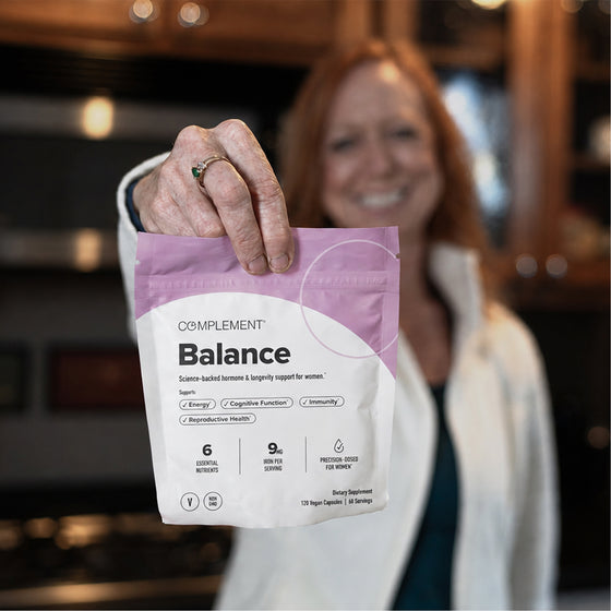 A smiling woman holds a Complement Balance supplement package toward the camera in a kitchen, with the product details visible and the background softly blurred.