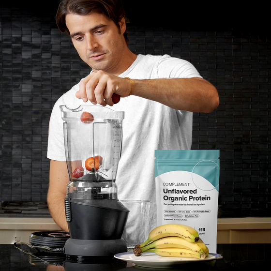 A person in a white shirt adds strawberries to a blender. A pack of Complement Unflavored Organic Protein and three bananas are on the nearby counter, set in a modern kitchen.