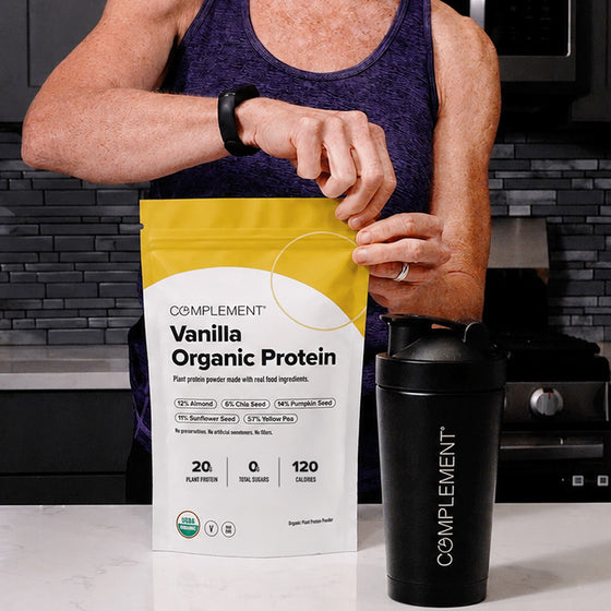 A person in a purple tank top opens a bag of Complement Vanilla Organic Protein powder in a kitchen, with a black Complement-branded shaker bottle on the counter.