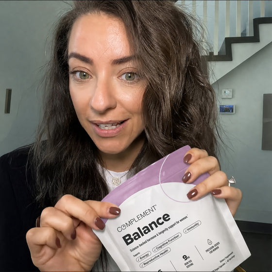 A woman with long brown hair holds a package labeled Complement Balance and smiles at the camera. A modern staircase and a light gray wall are visible in the background.