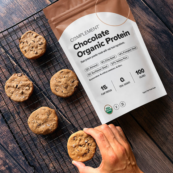 A hand reaches for a chocolate chip cookie on a cooling rack next to a bag of Complement Chocolate Organic Protein powder on a wooden table.