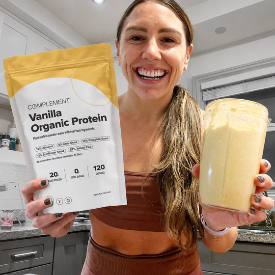 A smiling woman in a kitchen holds a packet of Complement Vanilla Organic Protein powder in one hand and a blender cup filled with a yellow smoothie in the other.
