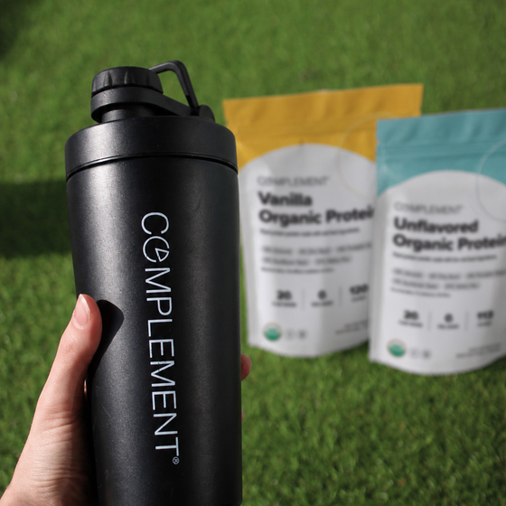 A hand holds a black COMPL✱MENT shaker bottle in front of two bags of Complement organic protein powder, one vanilla and one unflavored, on green grass.