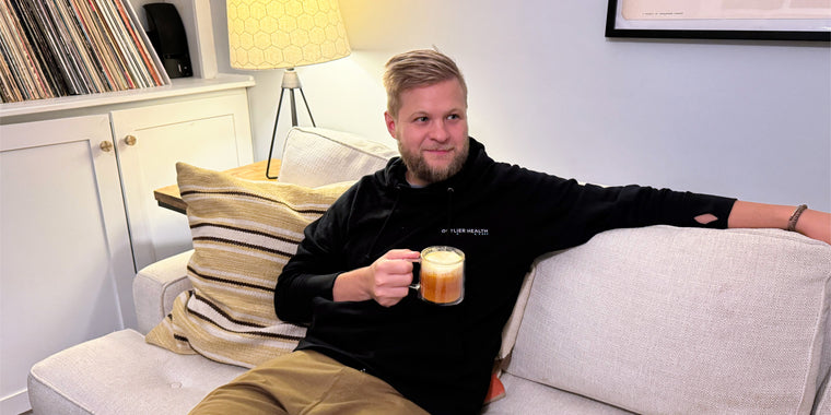 A man with short blond hair sits on a light-colored sofa, holding a mug of coffee. He wears a black hoodie and brown pants, and rests his arm on the back of the sofa. A lamp and records are visible in the background.