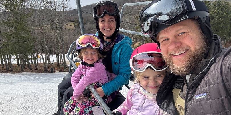 A family of four, wearing ski helmets and jackets, smiles while sitting on a ski lift surrounded by snow and trees on a winter day.