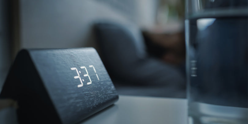 A digital clock on a bedside table displays the time 3:37. A glass of water and a blurred person lying in bed are visible in the background.